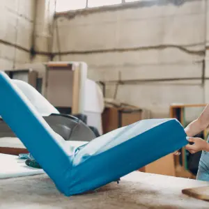 A craftsman working on a blue cushion cover in a professional workshop, illustrating expert upholstery techniques for restoring and customizing furniture. Image titled 'Types of Furniture We Upholster' highlights precision and quality in upholstery services.